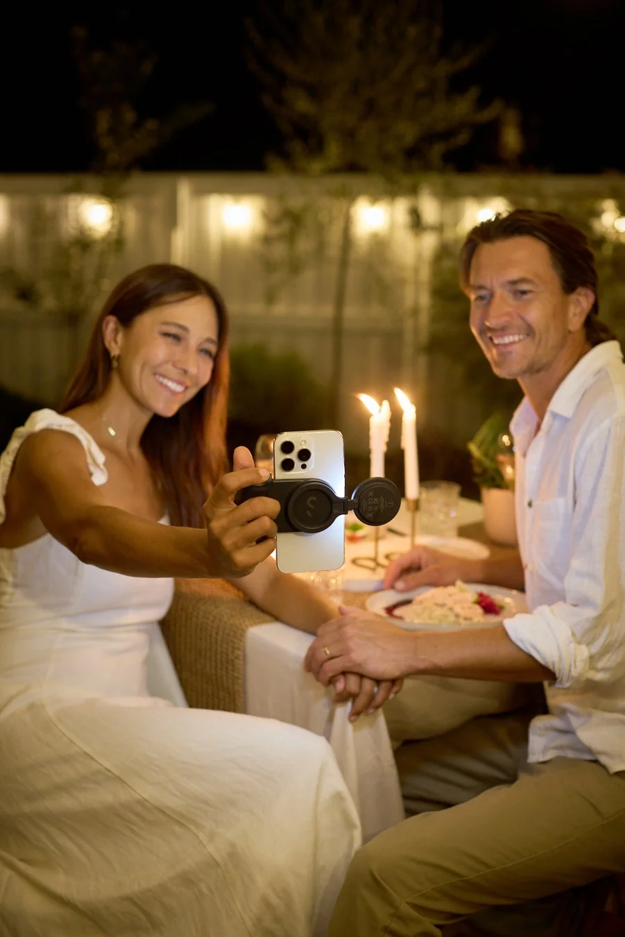 夜の屋外で、笑顔のカップルがキャンドルが灯るテーブルで食事をしながら、女性がスマートフォンで自撮りをしているロマンチックなシーンです。