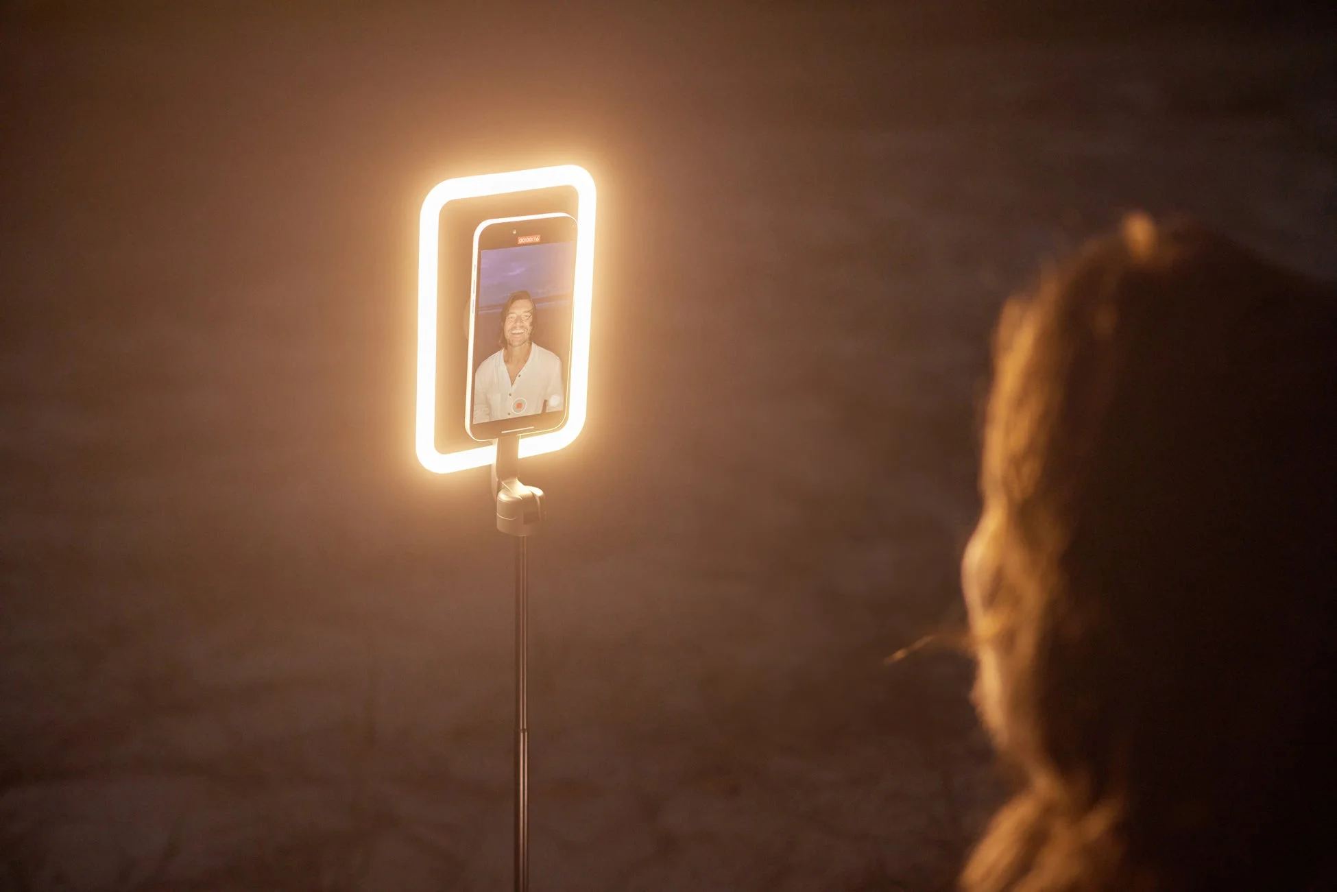 暗い場所でリングライト付きスマートフォンが設置され、画面には笑顔の男性が映っています。