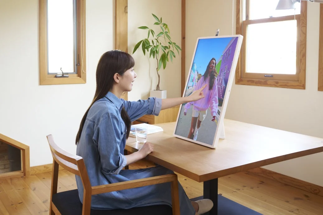部屋で女性が大きなディスプレイに映る女性の画像に触れている様子。リラックスした雰囲気の中でデジタルデバイスを操作している日常のワンシーンです。
