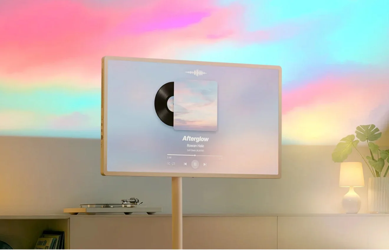 パステルカラーの美しい背景を映し出す壁の前に設置された、スタンド付きの薄型テレビが映し出されています。テレビ画面には「Afterglow」という曲のアルバムアートが表示されており、その下にはターンテーブルが置かれています。部屋は柔らかな光に包まれ、観葉植物やランプが落ち着いた雰囲気を醸し出しています。