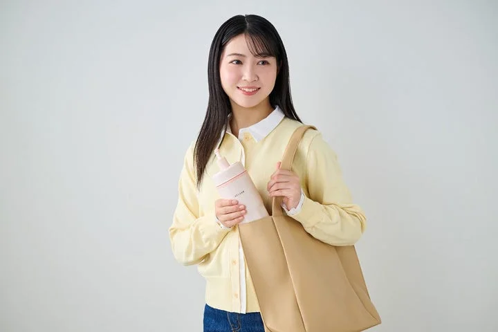笑顔の若い女性が、ピンク色のボトルをベージュのトートバッグに入れている様子