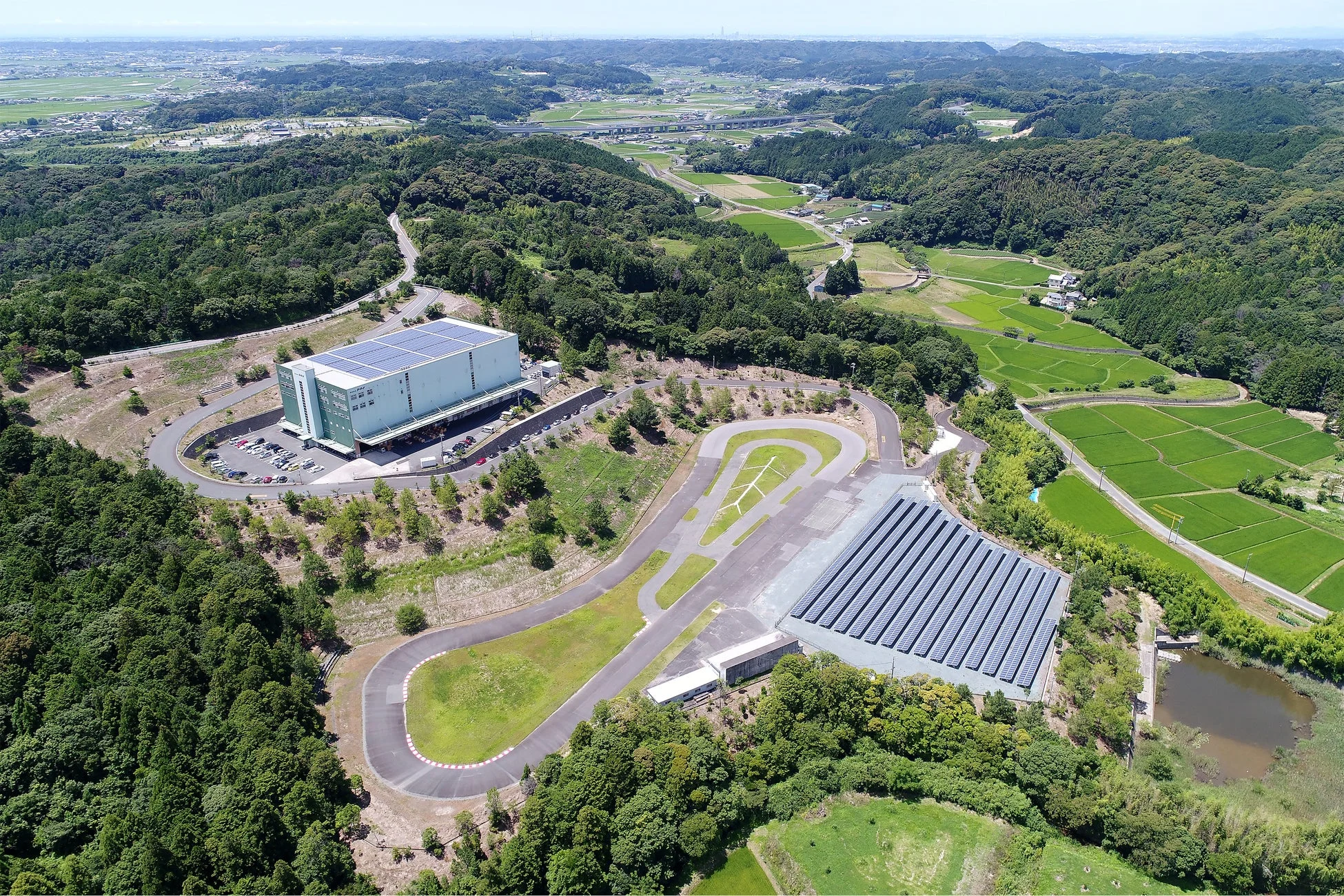 空撮された広大な風景で、屋根にソーラーパネルを設置した大きな倉庫と、隣接するテストコース、そして大規模な太陽光発電所が見えます。周囲は豊かな森林と水田に囲まれ、自然と産業が共存する郊外の景色が広がっています。